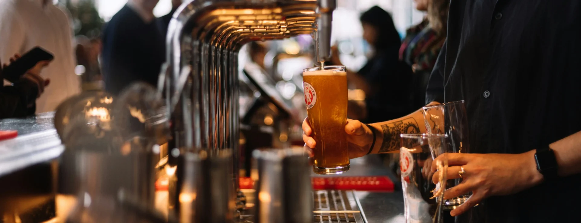 A bartender hands a freshly poured pint of beer across the bar, with taps and customers in the background
