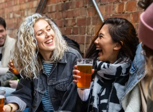 Friends laughing together outdoors while holding pints of beer at a brick-walled pub