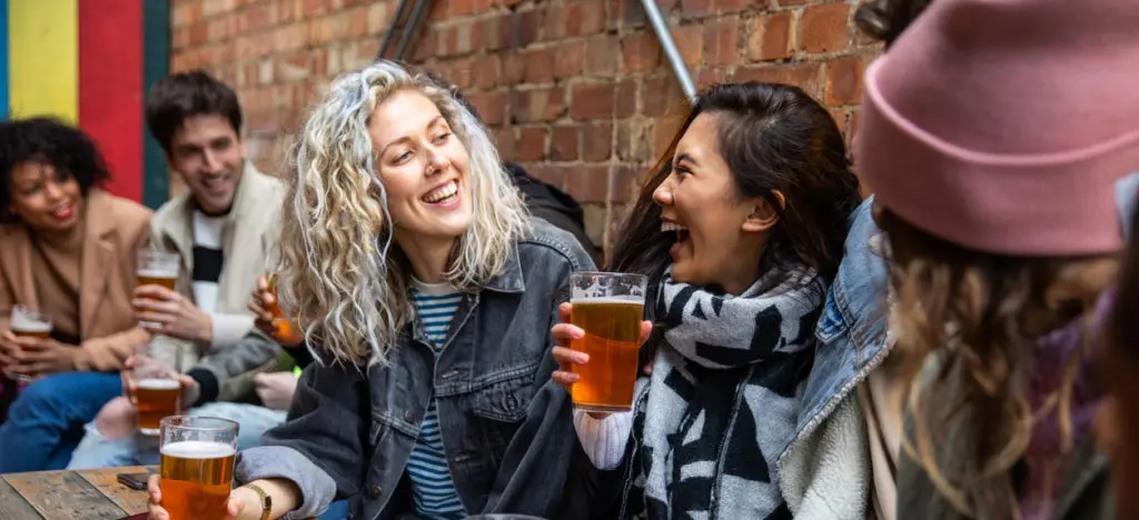 Friends laughing together outdoors while holding pints of beer at a brick-walled pub