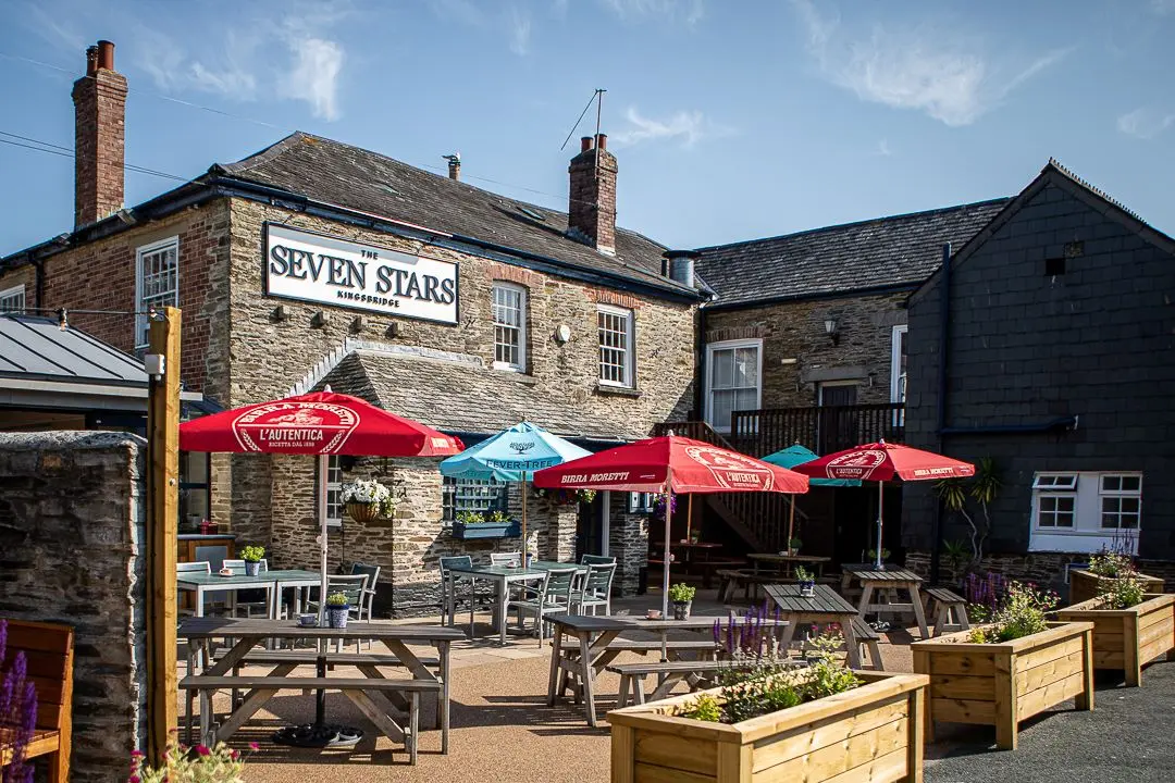 Stone building with sign for The Seven Stars pub, outdoor seating area with tables and coloured umbrellas