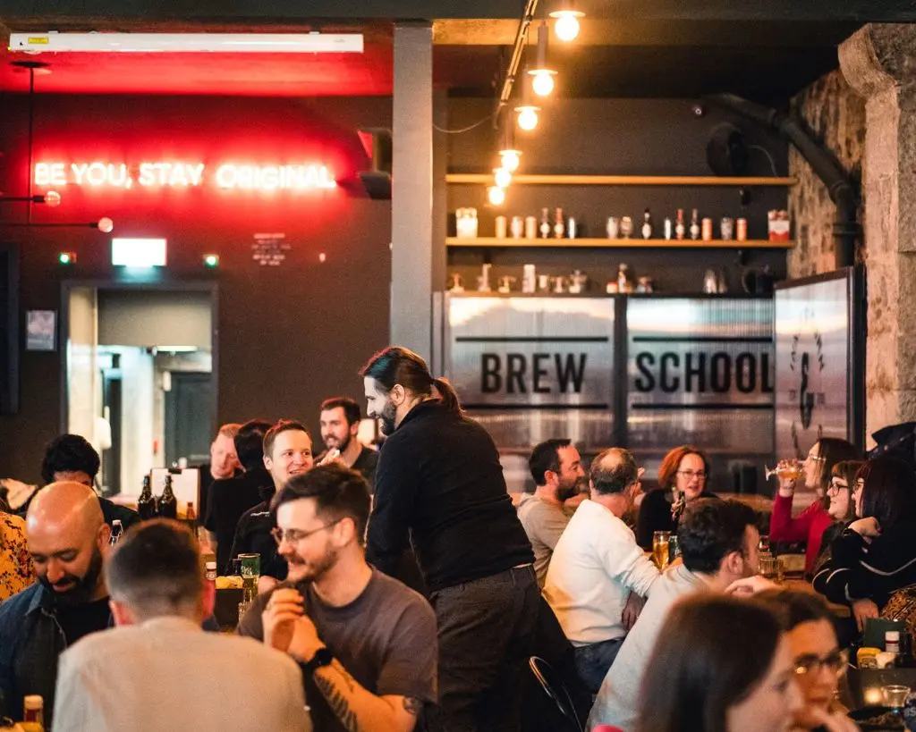 People gathered with drinks at Brew School bar, under a neon sign reading "Be You, Stay Original