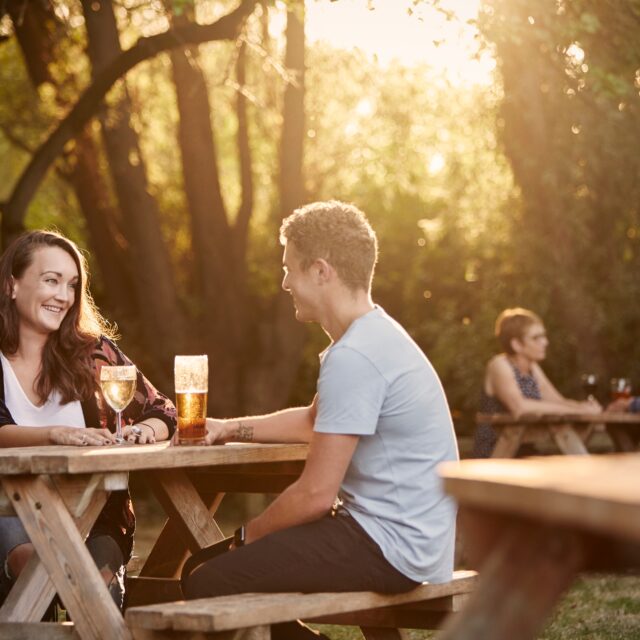 People sitting at wooden picnic tables outdoors, drinking wine and beer in sunlit woodland setting