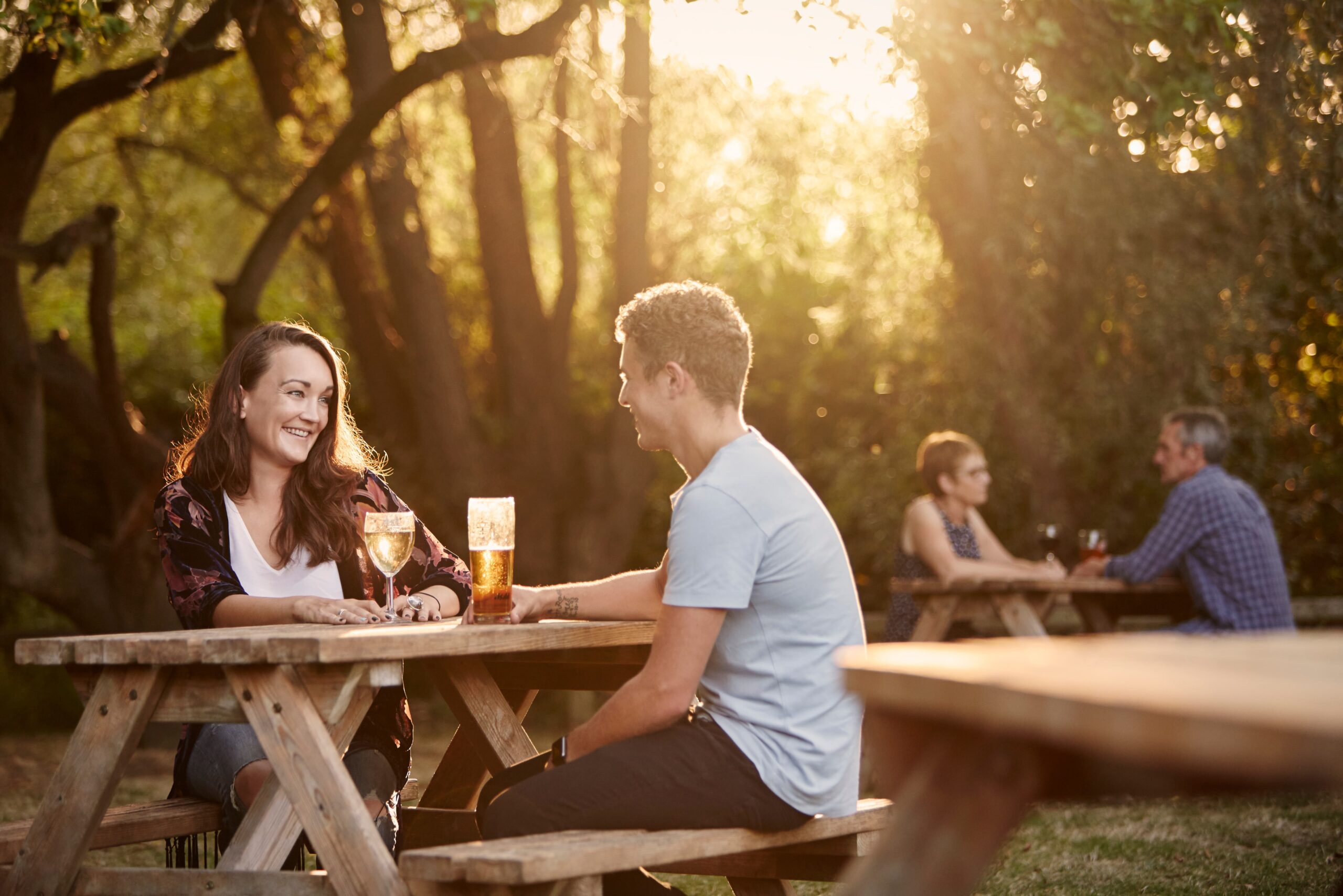 People sitting at wooden picnic tables outdoors, drinking wine and beer in sunlit woodland setting
