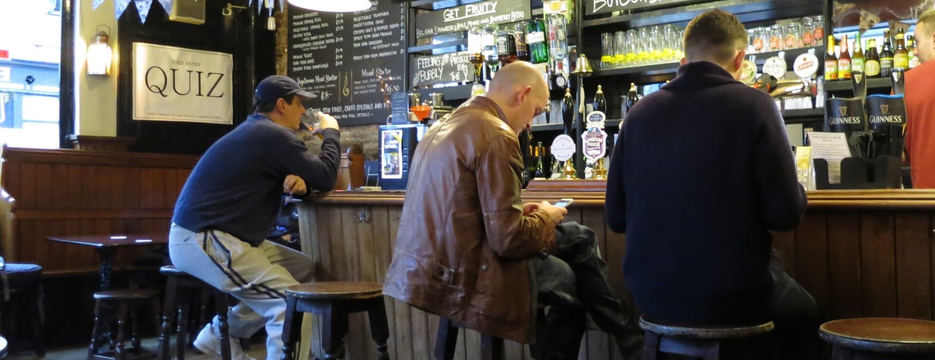 Men sitting on stools at a pub bar, with chalkboard drinks menus and a Free Entry Quiz sign displayed nearby