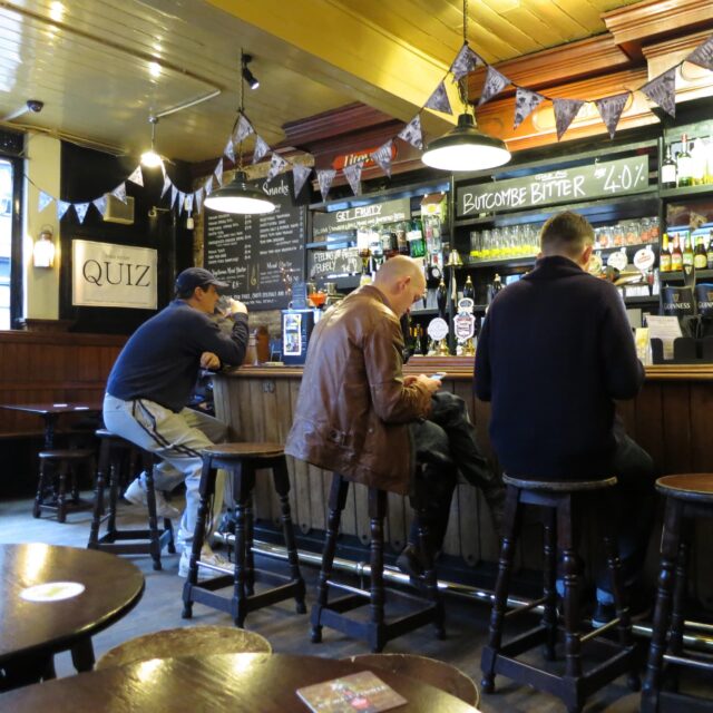 Men sitting on stools at a pub bar, with chalkboard drinks menus and a Free Entry Quiz sign displayed nearby