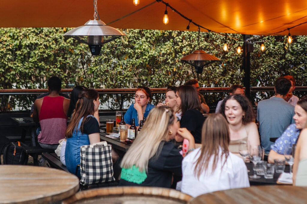 People chatting over drinks at a busy outdoor pub beer garden with hedges and string lights