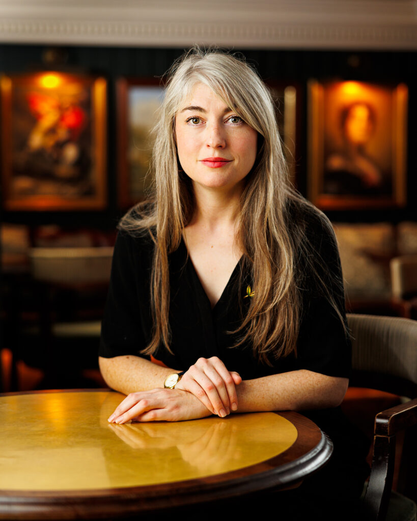 Woman with long grey hair in a black top sits with hands folded on a table in a wood-panelled room with framed artwork