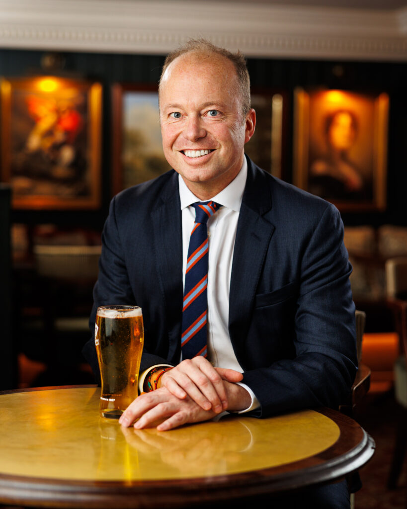 Man in a navy suit and striped tie smiling at a pub table with a pint of beer