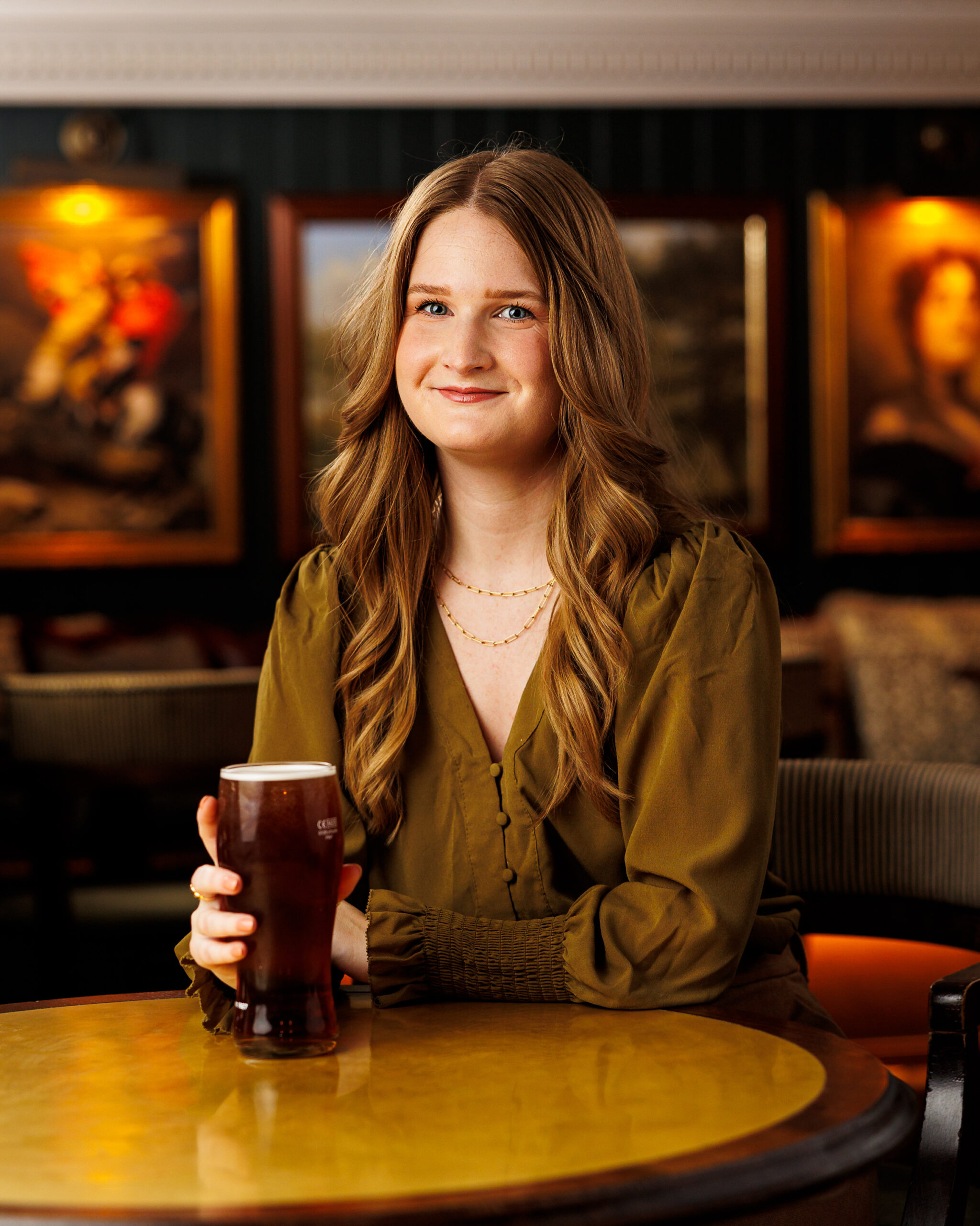Woman with long wavy hair smiling in a pub, holding a pint of ale at a table