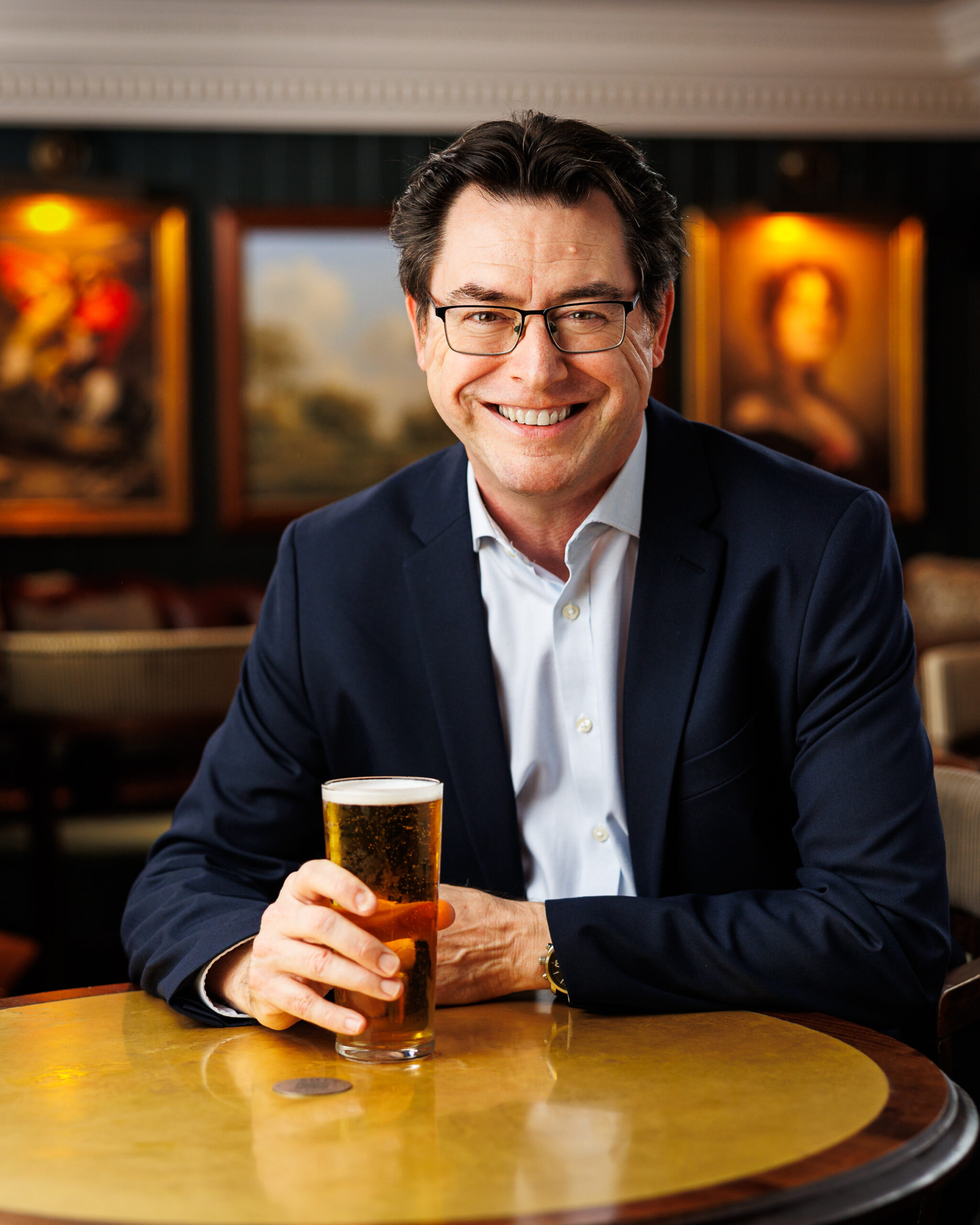 Man in glasses and navy suit smiling while holding a pint of beer at a pub table