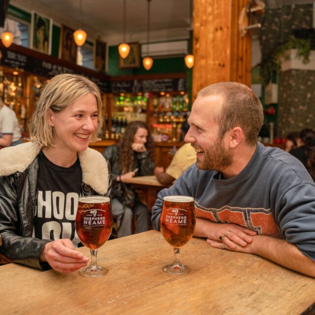 A man and woman laugh together over pints of Shepherd Neame ale at a busy pub table
