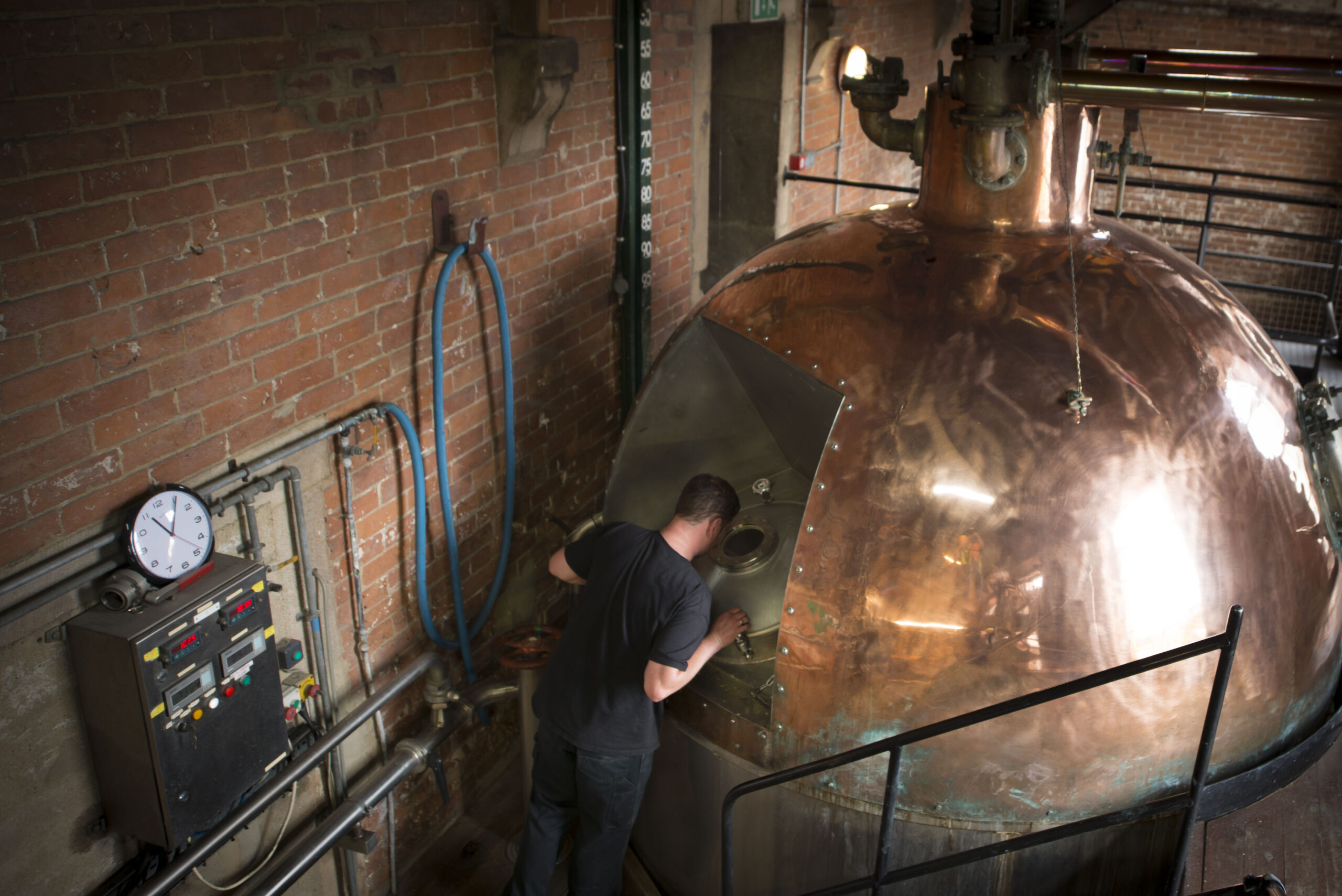 A worker inspects the hatch of a large copper still in a distillery, surrounded by pipework and brick walls