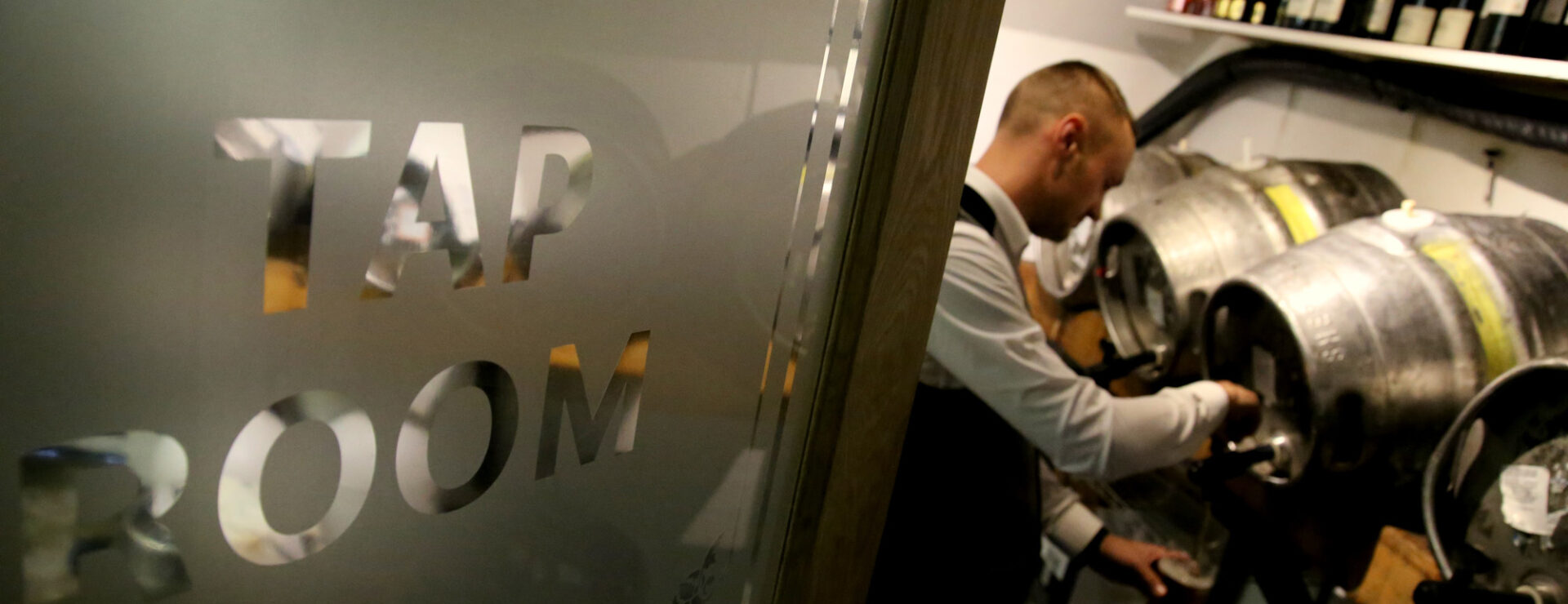 A member of staff pours a pint from a cask through the "Tap Room" doorway, with wine bottles stored above