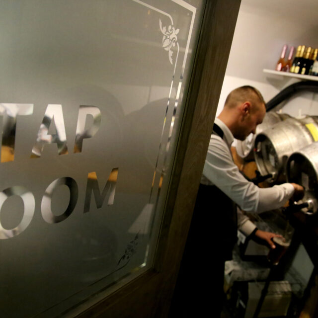 A member of staff pours a pint from a cask through the "Tap Room" doorway, with wine bottles stored above