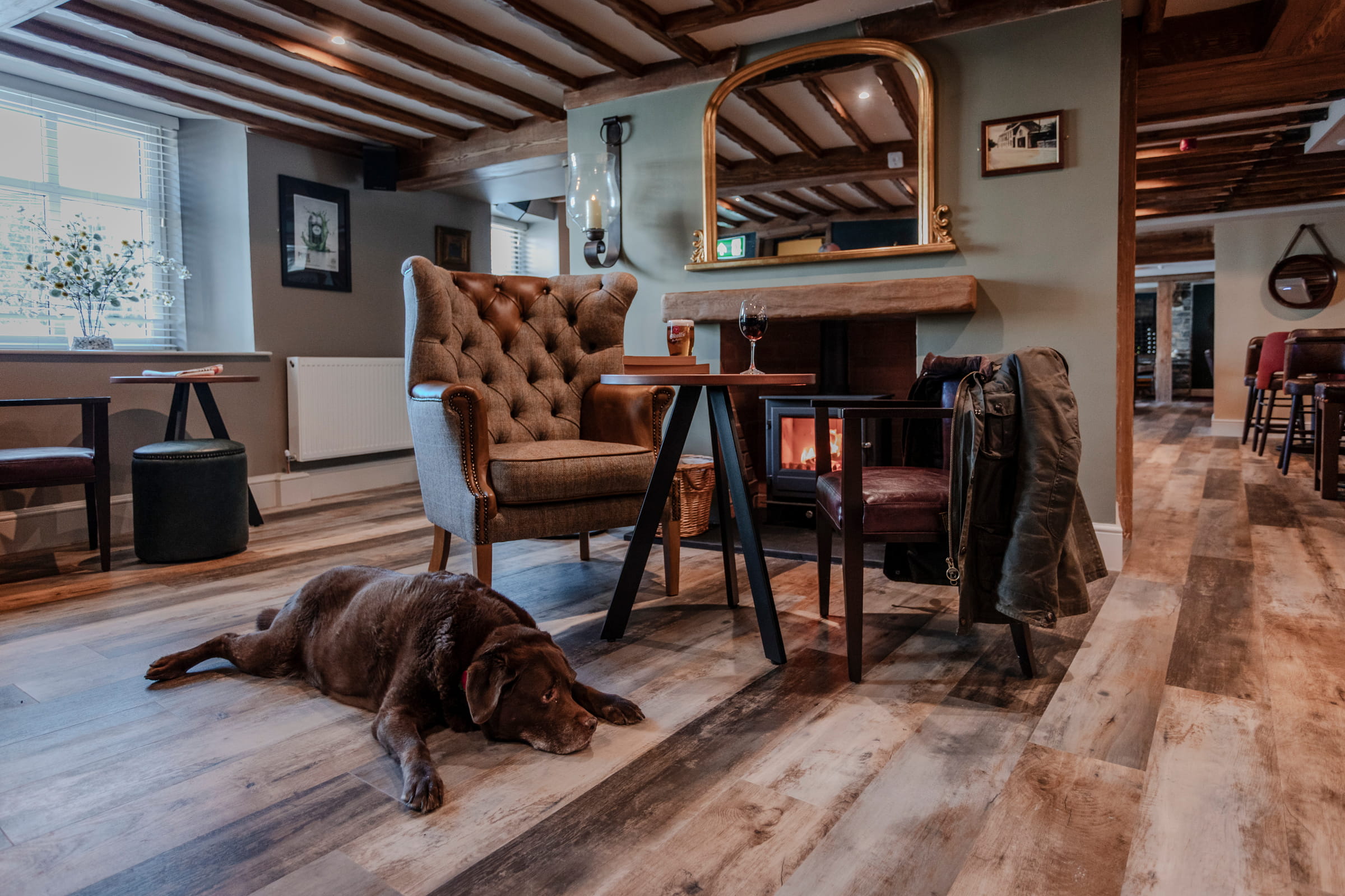 A chocolate Labrador lies by the fireside in a cosy pub lounge with beamed ceilings and a lit wood burner