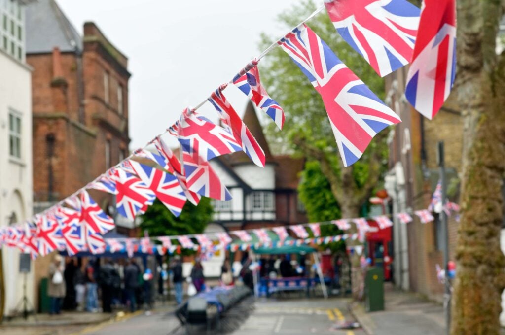 Union Jack bunting strung across a street lined with old buildings, with market stalls and people gathered below