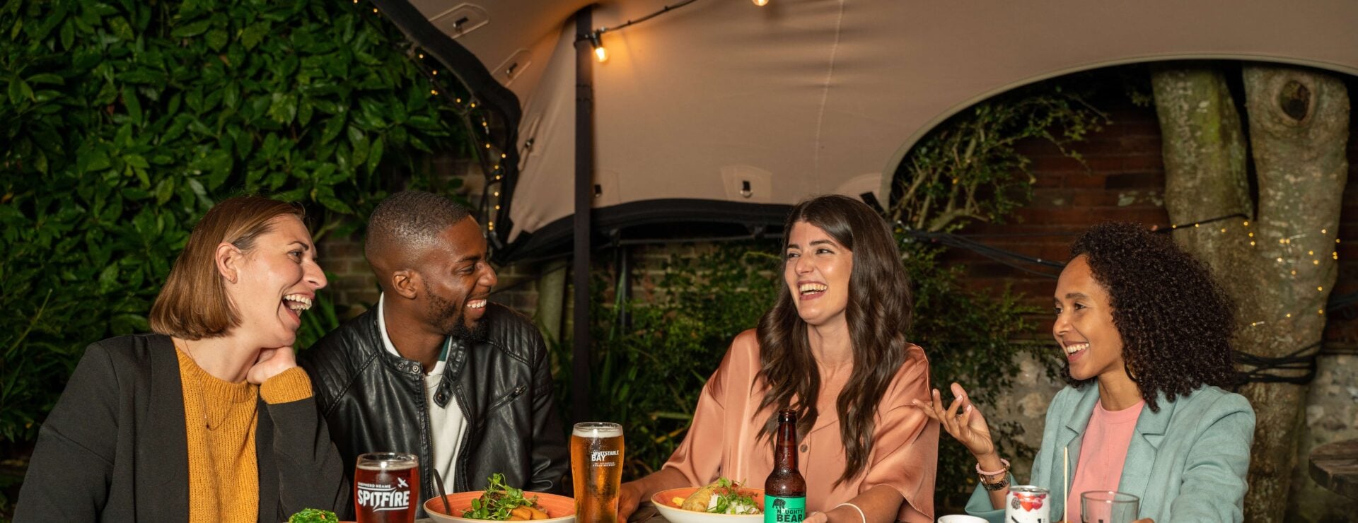 Four friends laughing together over drinks and food at a wooden table in a covered pub garden area
