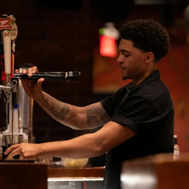 A bartender with a tattooed arm pours a drink from a tap into a glass at a dimly lit bar