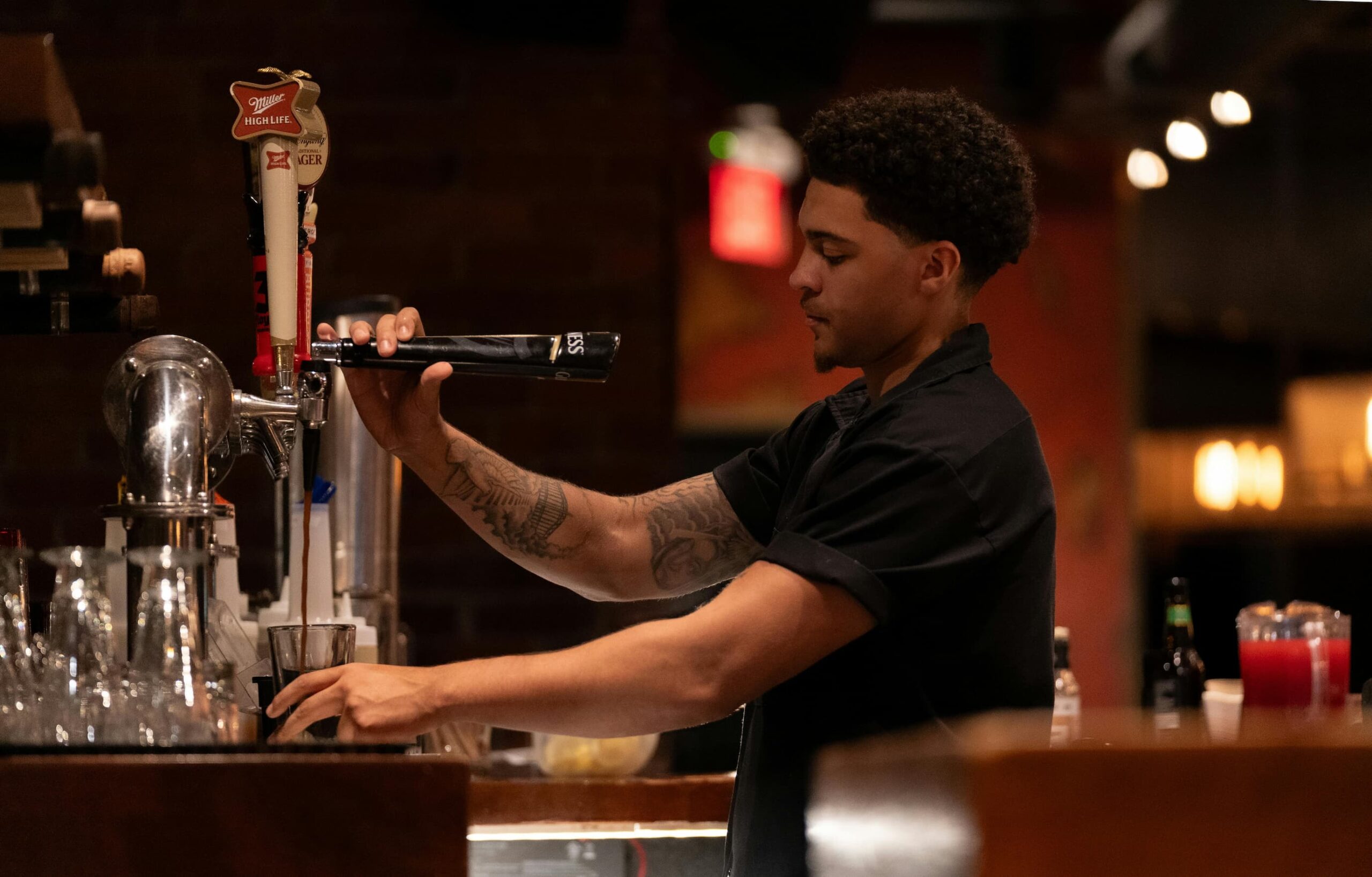 A bartender with a tattooed arm pours a drink from a tap into a glass at a dimly lit bar