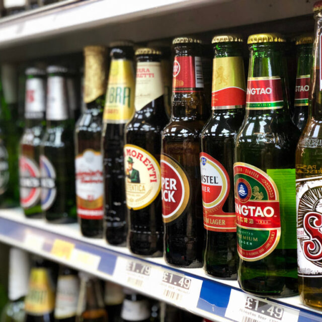 Row of international beer bottles on a shop shelf, including Moretti, Tsingtao and Sol, with price labels below
