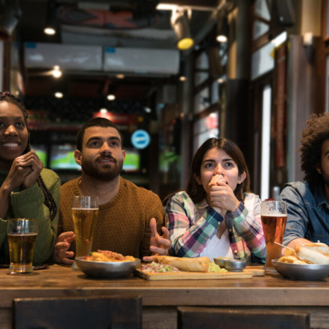 Four friends watch a match intently at a pub table with beers and snacks, showing tense expressions