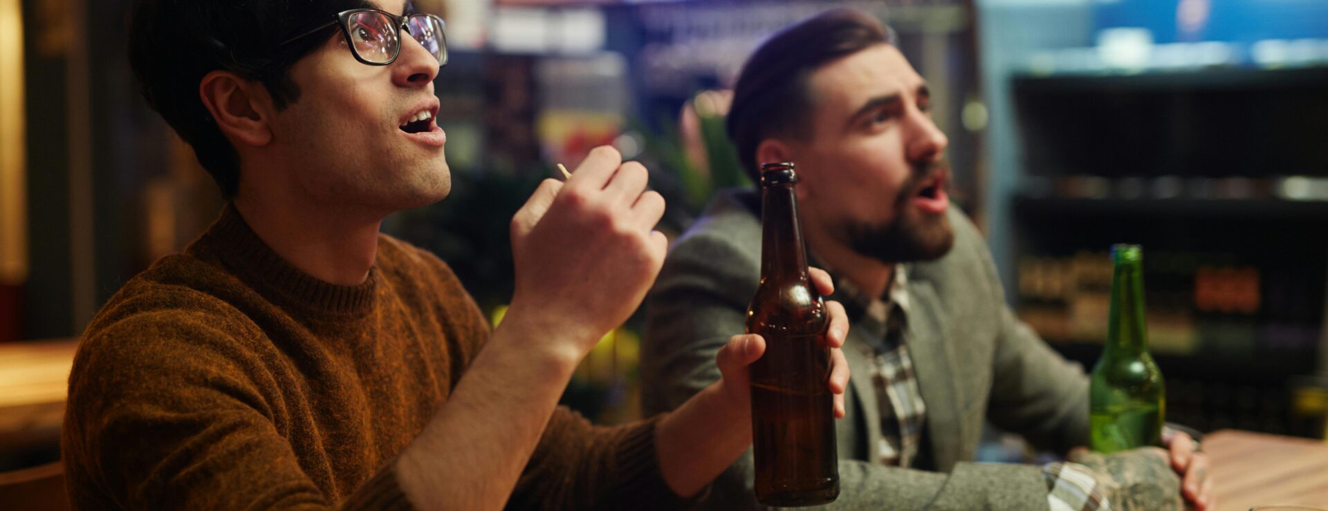 Two men at a bar with beer bottles and snacks, reacting excitedly while watching a screen out of view