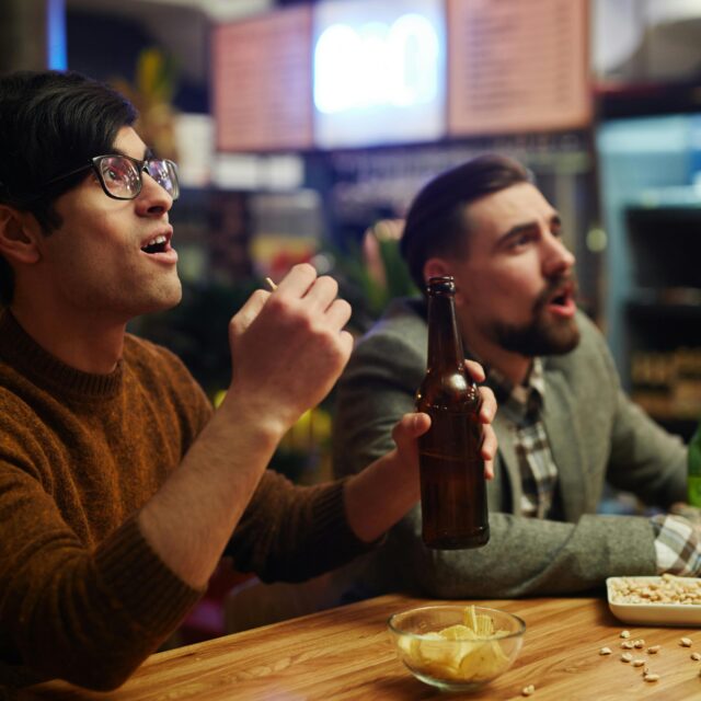 Two men at a bar with beer bottles and snacks, reacting excitedly while watching a screen out of view
