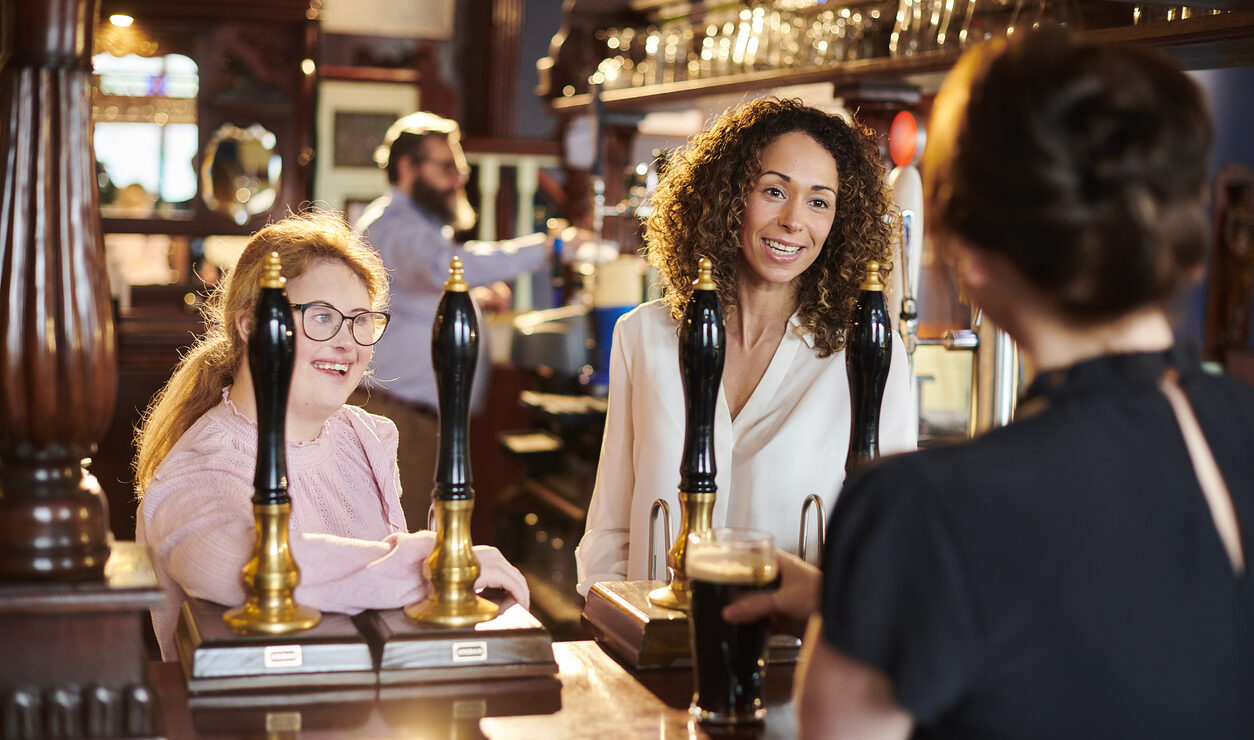 Two women at a pub bar being served a pint by a bartender, with a man working behind the bar