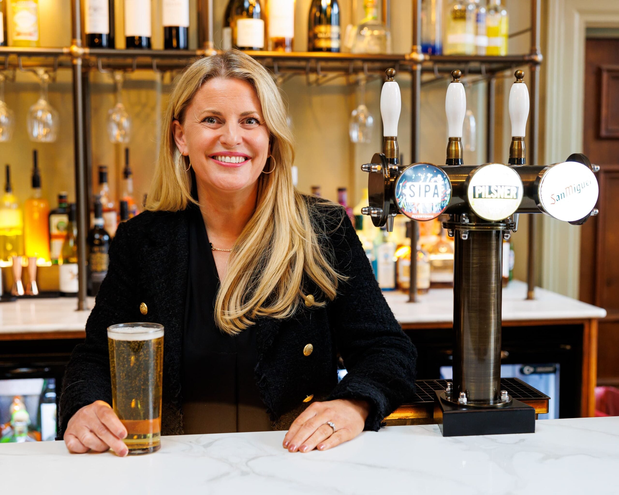 Smiling woman standing behind a bar with a pint of lager, next to beer taps for SIPA, Pilsner and San Miguel