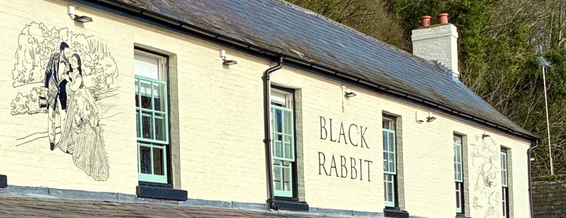 The Black Rabbit pub, a cream-painted building with a covered veranda, picnic tables and hanging flower baskets, set against trees