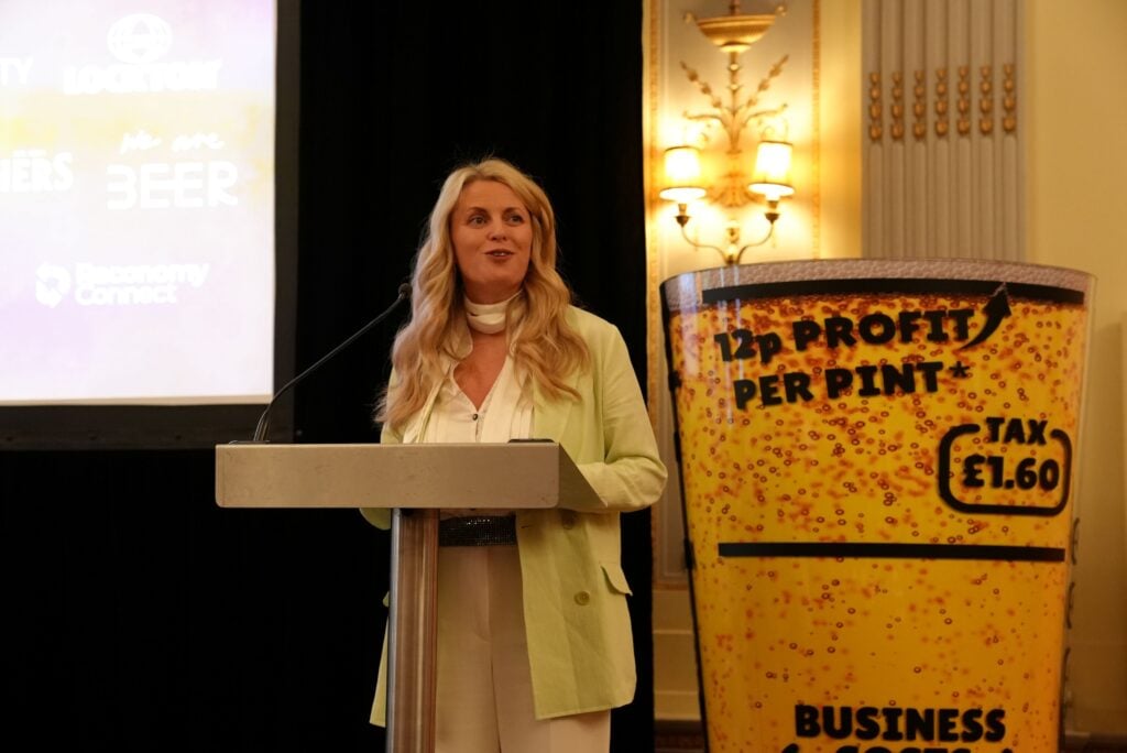Speaker at a podium beside a beer pint-shaped sign reading "12p profit per pint, tax £1.60