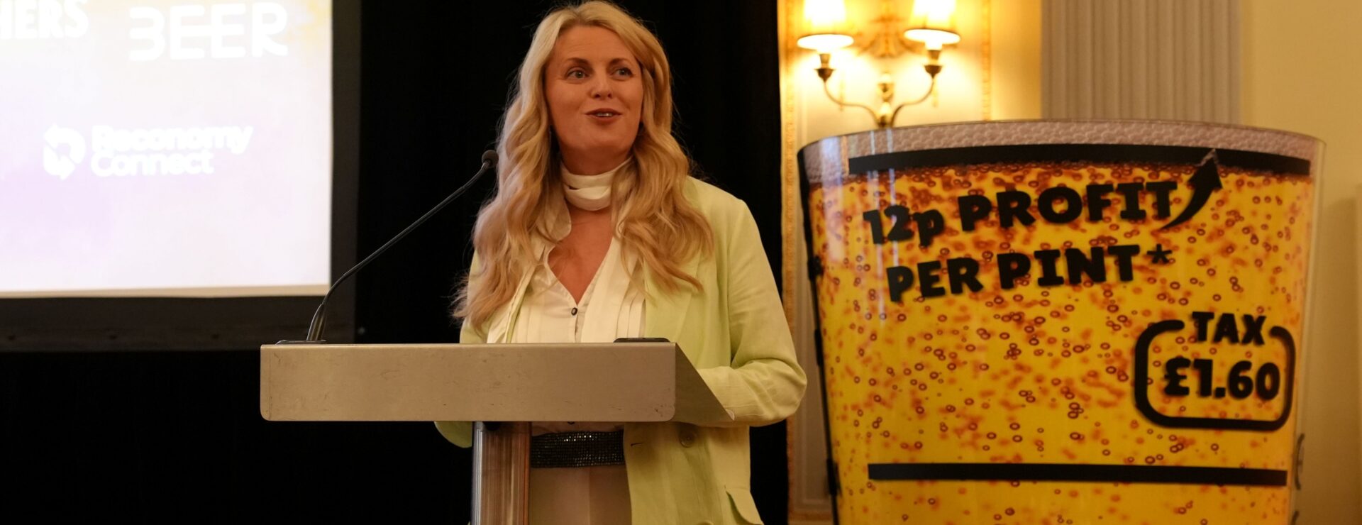 Speaker at a podium beside a beer pint-shaped sign reading "12p profit per pint, tax £1.60