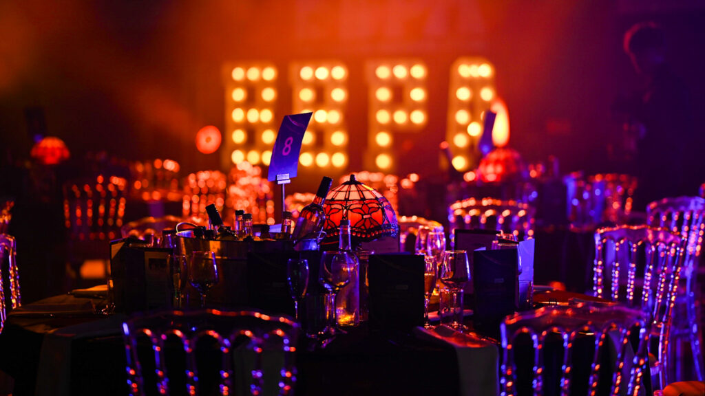 Gala dinner table set with wine glasses and a lamp, lit by a large illuminated marquee sign in the background