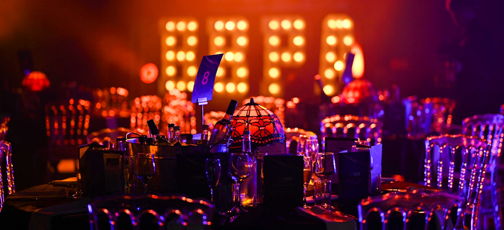 Gala dinner table set with wine glasses and a lamp, lit by a large illuminated marquee sign in the background